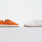 Jägermeister x Flowers For Society The Curt Sneaker, båda färgvarianter sida vid sida — orange mocka och vitt läder — på vit piedestal