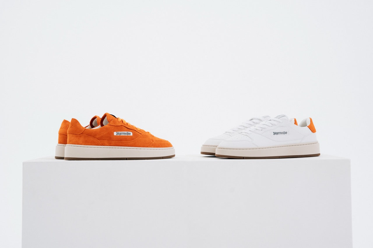 Jägermeister x Flowers For Society The Curt Sneaker, båda färgvarianter sida vid sida — orange mocka och vitt läder — på vit piedestal