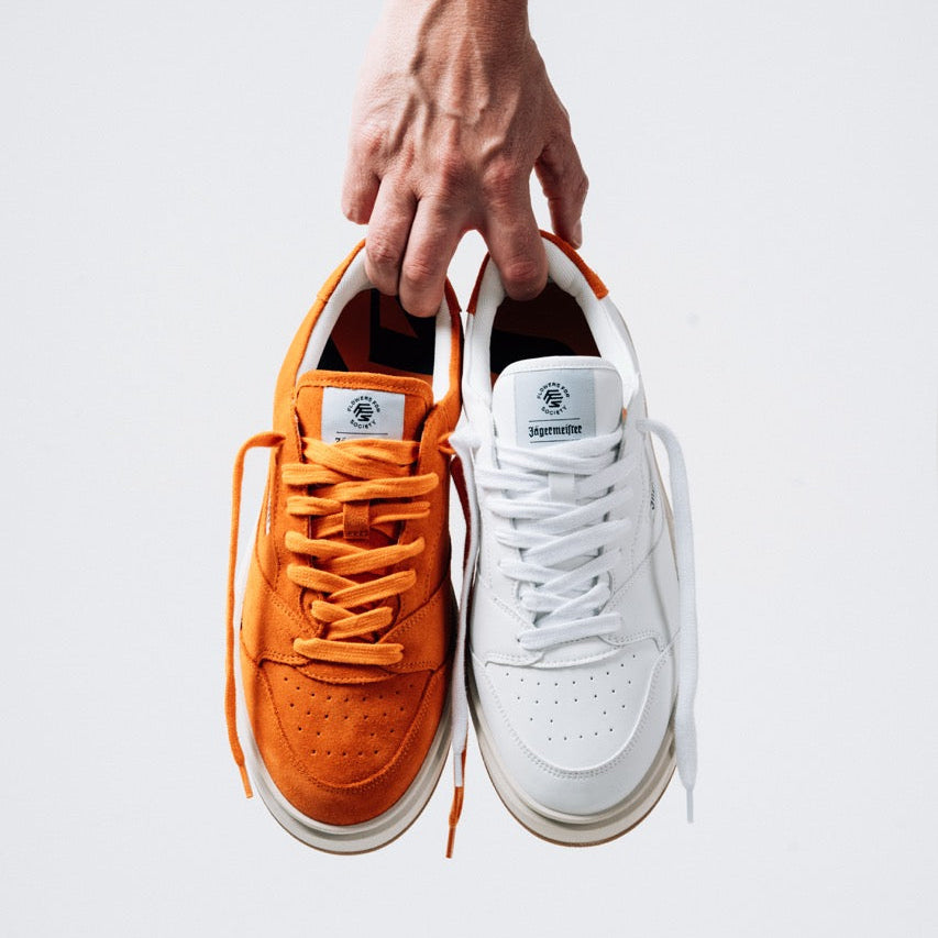 Jägermeister x Flowers For Society The Curt Sneaker, hand håller upp båda färgvarianter i orange mocka och vitt läder framifrån