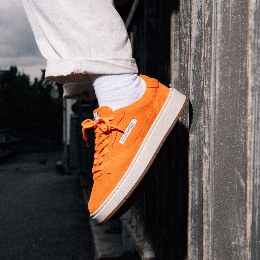 Jägermeister x Flowers For Society The Curt Sneaker i orange mocka, sidovy på fot mot vägg utomhus